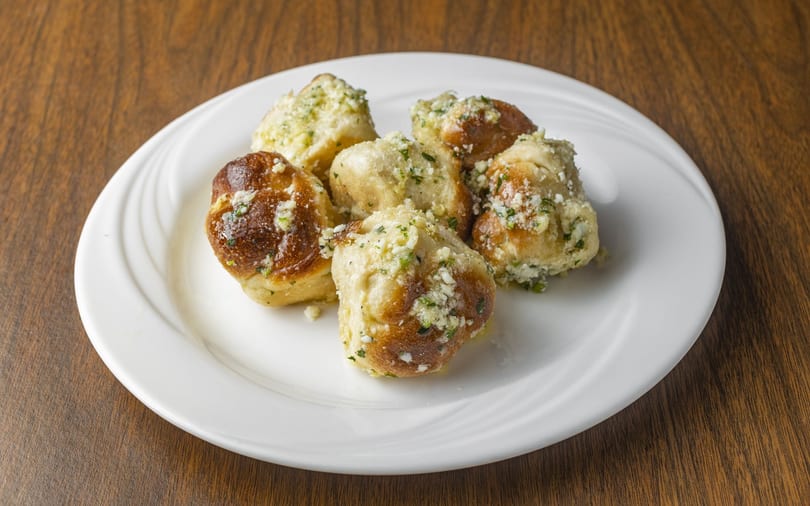 Six golden-brown garlic knots with herbs and cheese on a white plate.