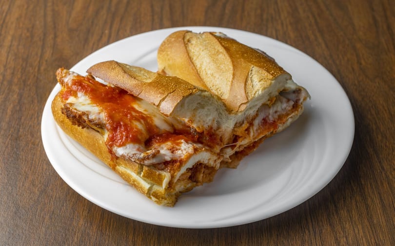 A chicken parmesan hero sandwich, cut in half, on a white plate on a wooden table.